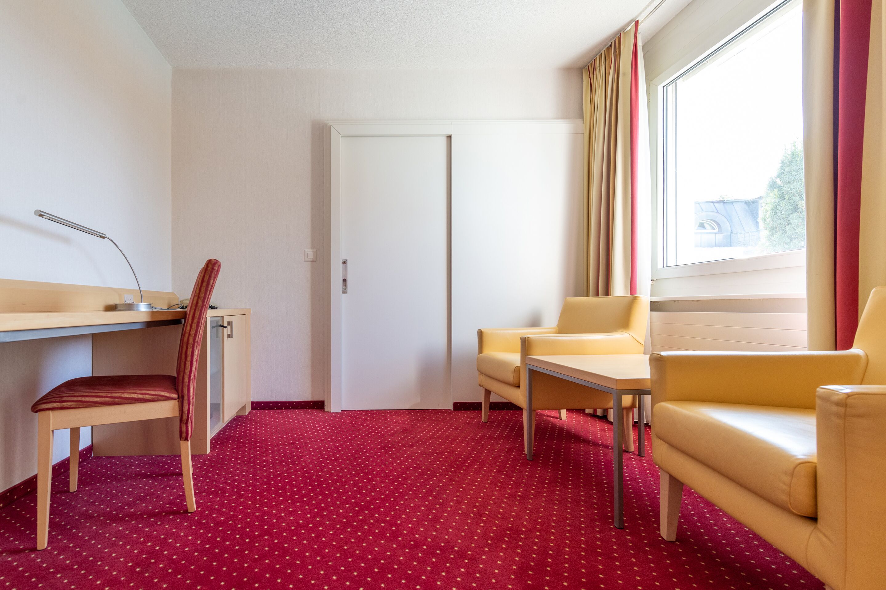 Habitación de hotel con escritorio, dos sillas, alfombra roja y ventana con cortinas.