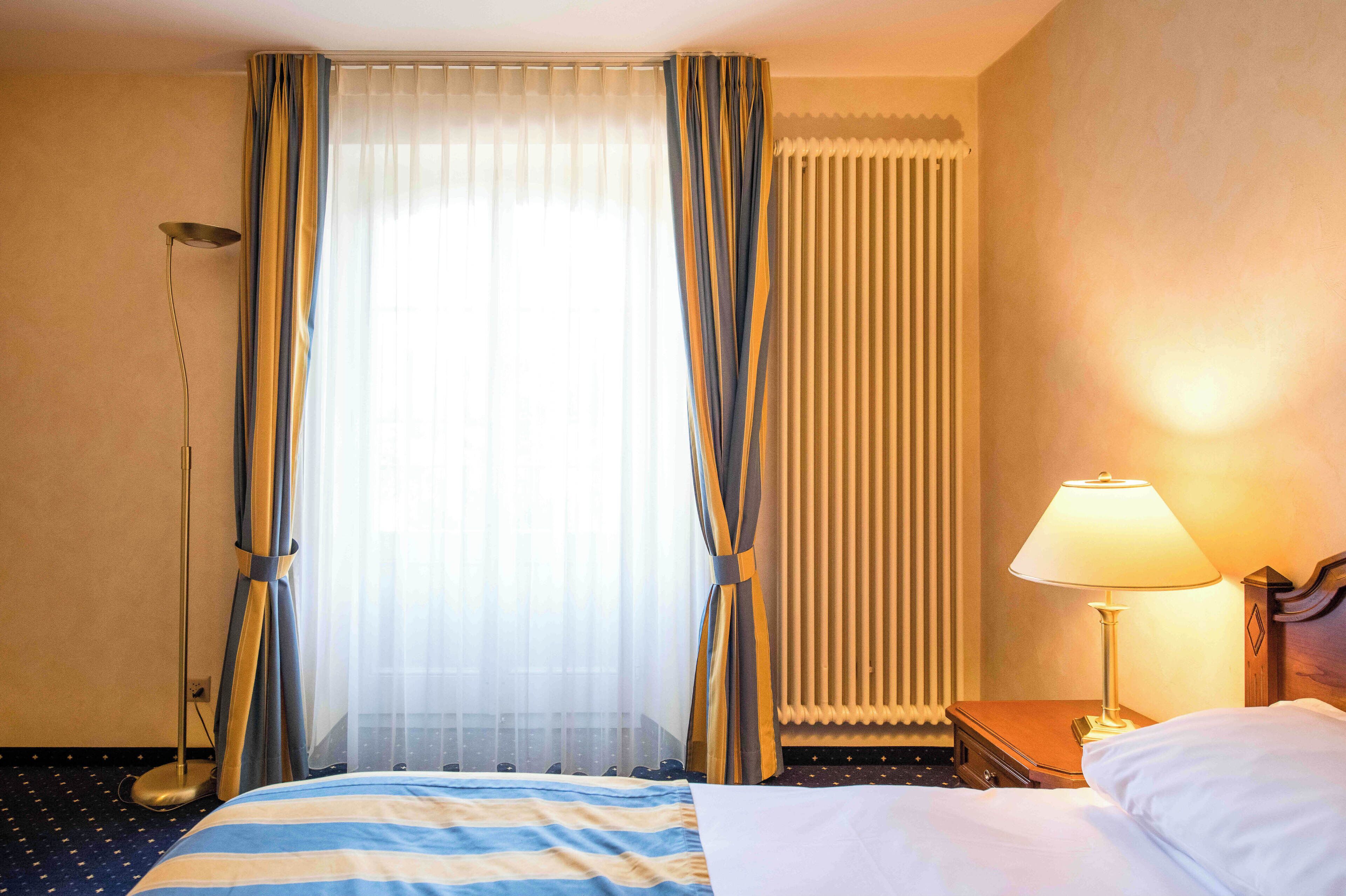 A hotel room with a bed, curtains, a floor lamp, a bedside table with a lamp, and a radiator.