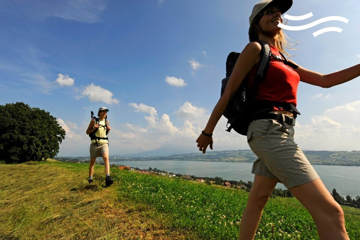 Eine Person wandert auf einer grasbewachsenen Anhöhe, mit Rucksack, mit Blick auf einen See und ferne Berge.