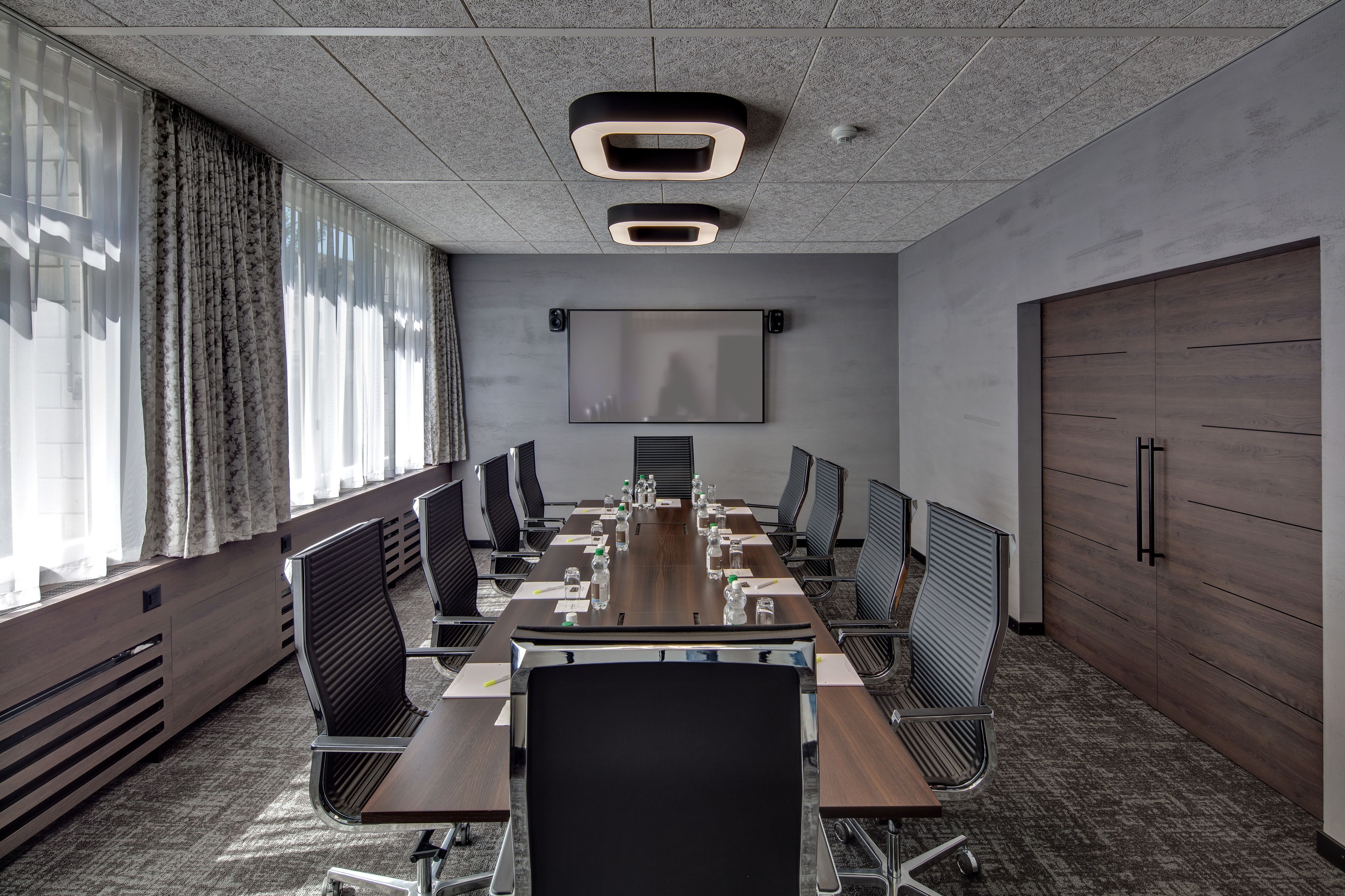 A conference room with a long table, chairs, a projector screen, and a large window with curtains.