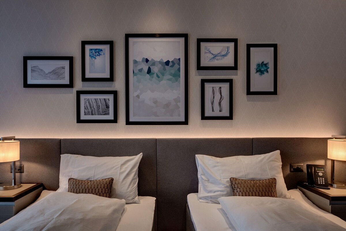 Habitació d'hotel amb dos llits, un capçal, taules de nit, làmpades i una paret decorada amb quadres.