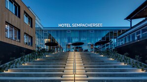 A modern hotel entrance with a glass facade, a staircase leading up to the entrance, and outdoor seating areas.