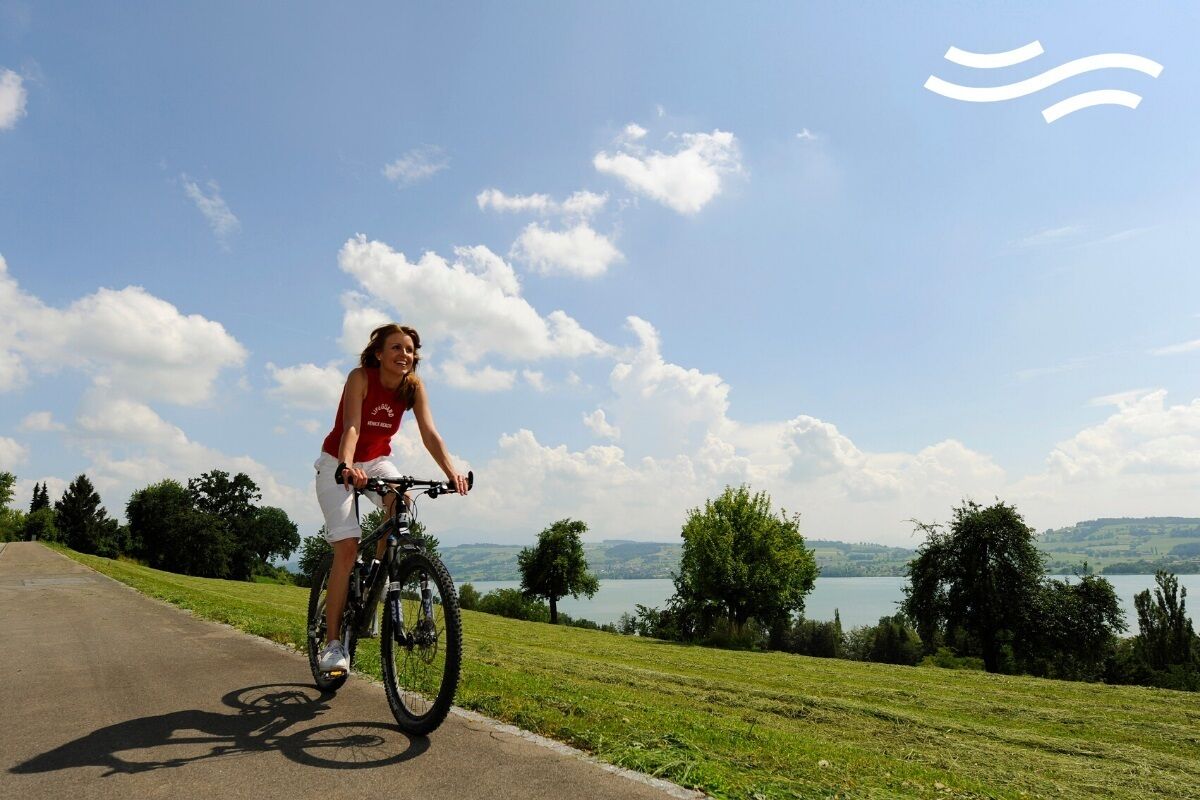 A person riding a bicycle on a paved path with a scenic view of a lake and hills in the background.