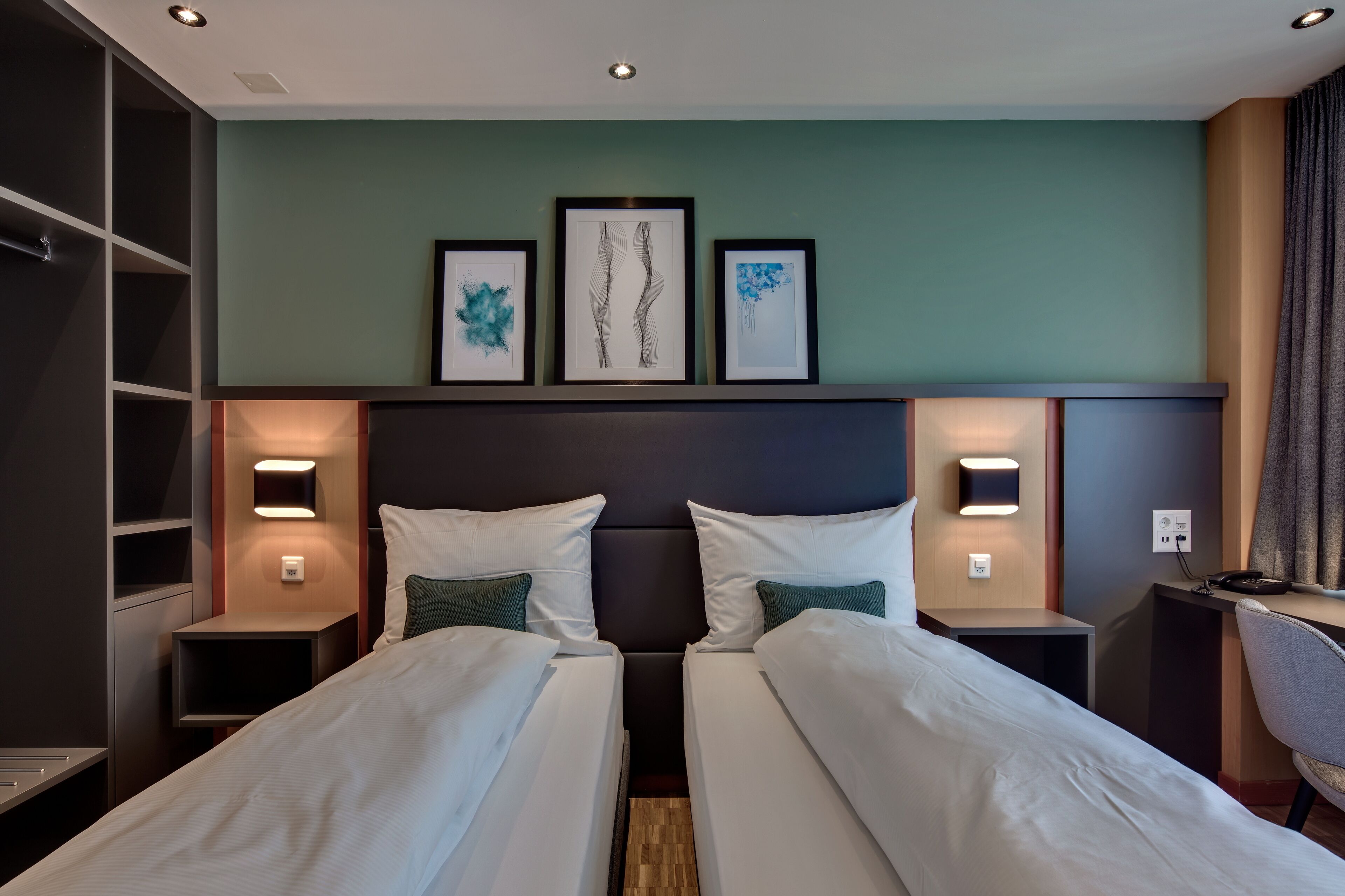 A hotel room with two beds, a desk, and a chair. There are wall-mounted lights and a shelf with framed artwork.