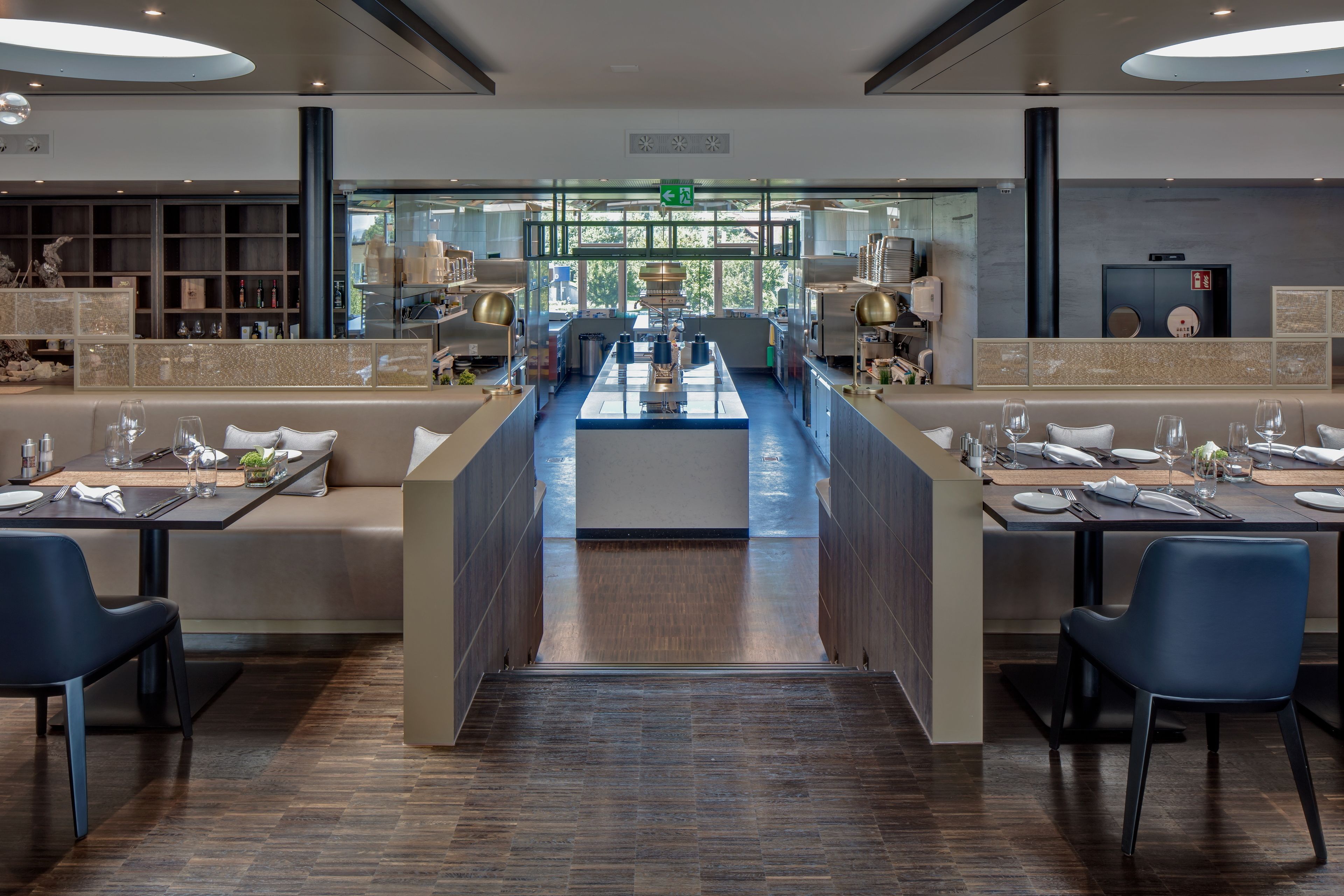 A modern restaurant with a central island, seating on both sides, and a view through large windows.