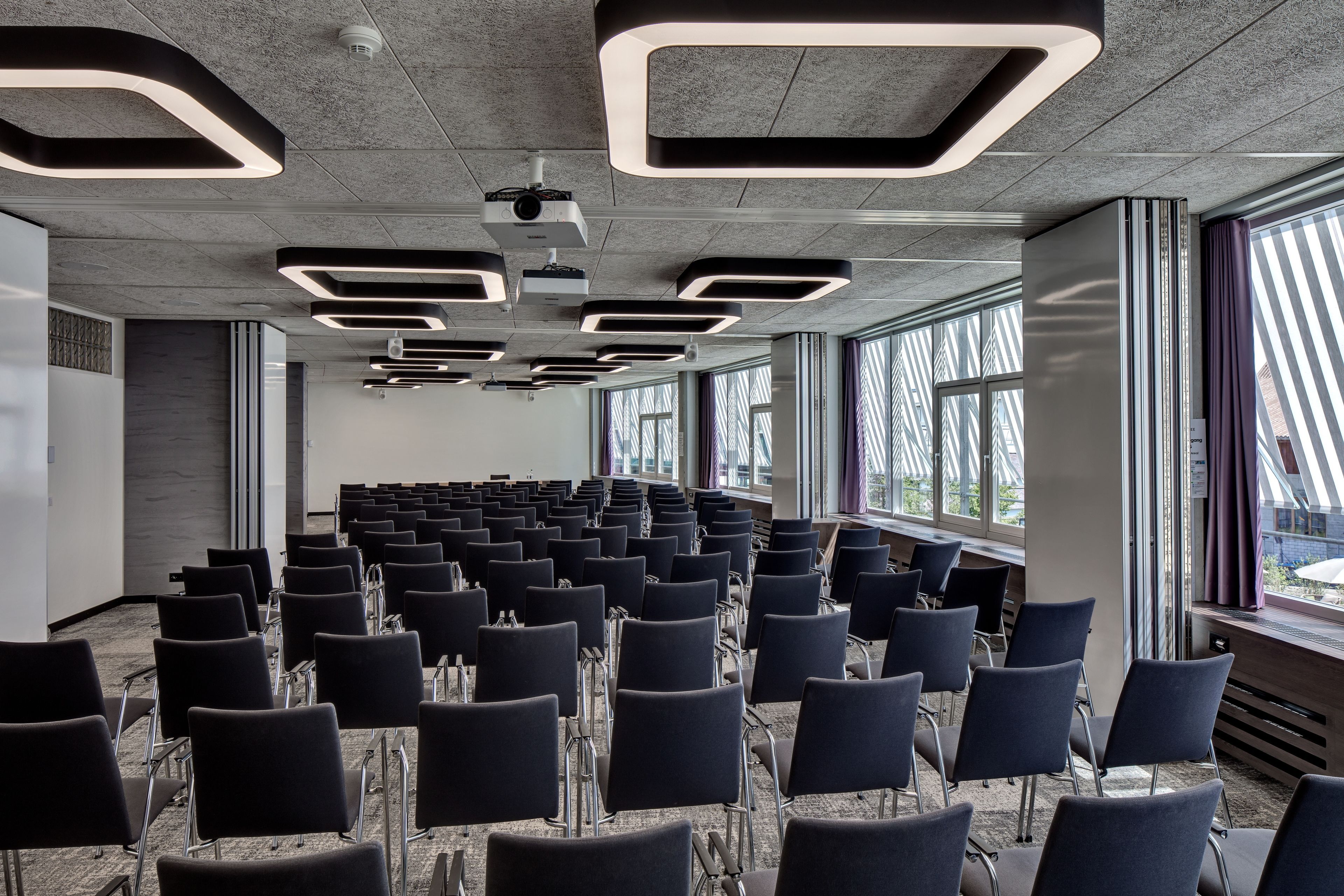 A conference room with rows of chairs, ceiling projectors, and large windows with blinds.