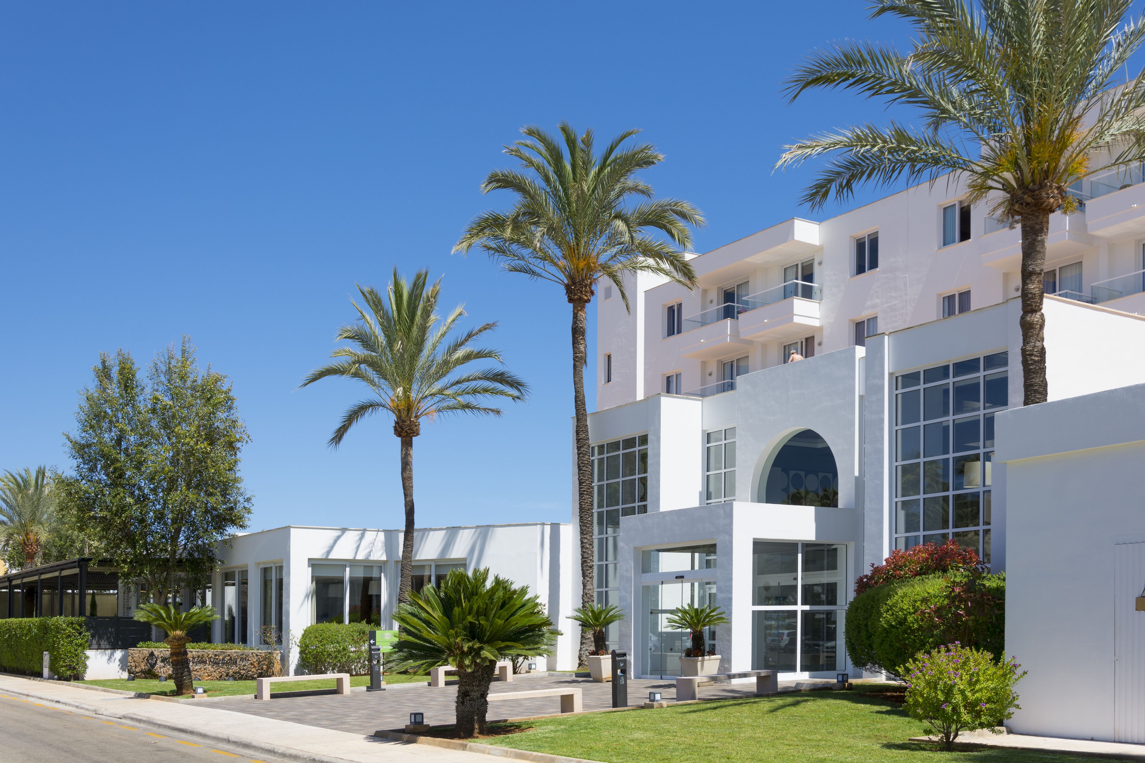 Un bâtiment moderne à étages, doté de grandes fenêtres et d’une pelouse bien entretenue agrémentée de palmiers.
