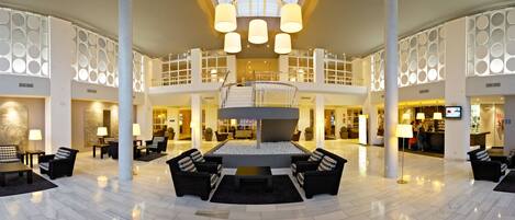 En rummelig lobby med et centralt siddeområde, flere etager og et moderne design med runde lysarmaturer.