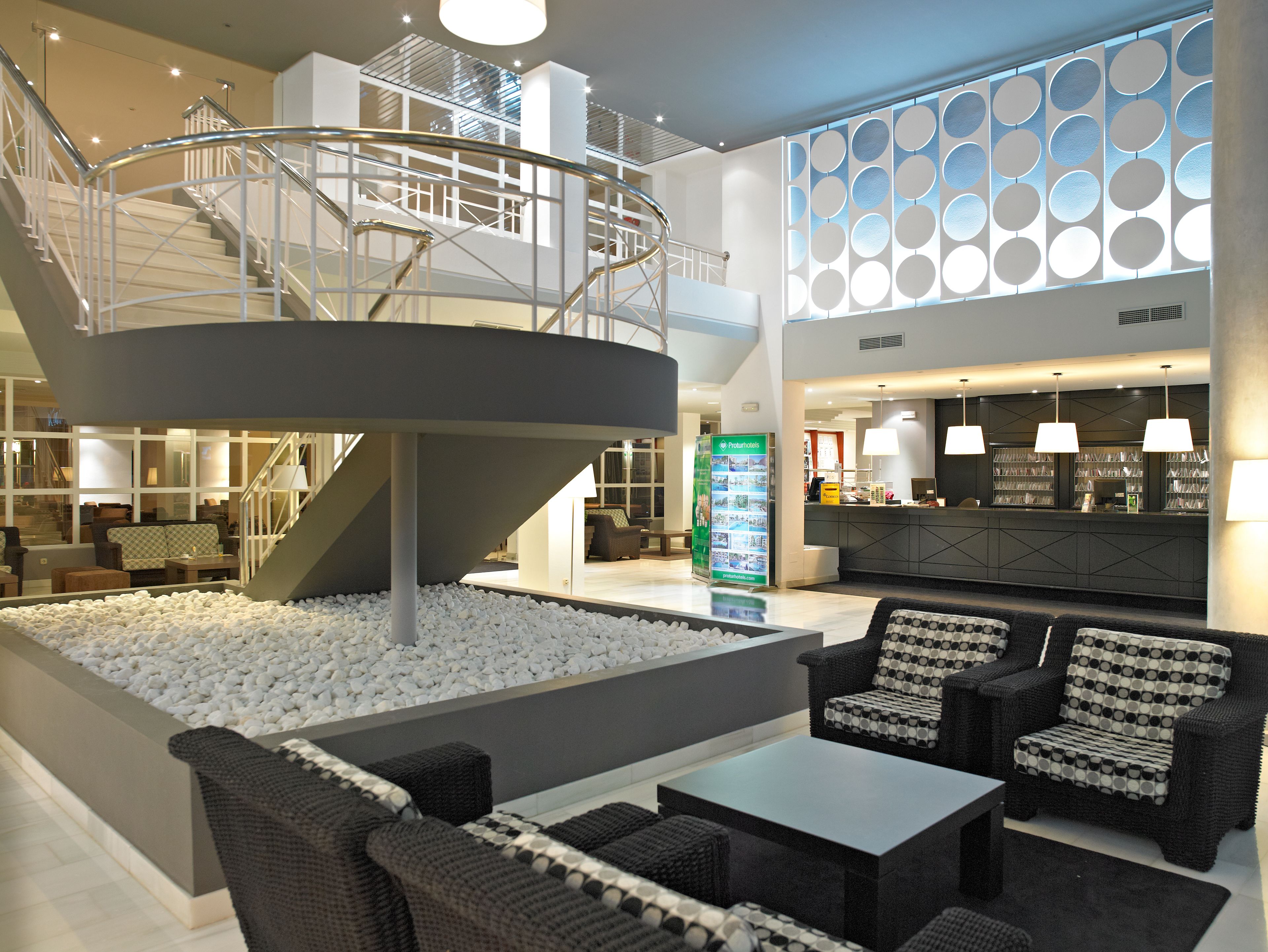A modern hotel lobby with a central seating area, a staircase, and a reception desk.