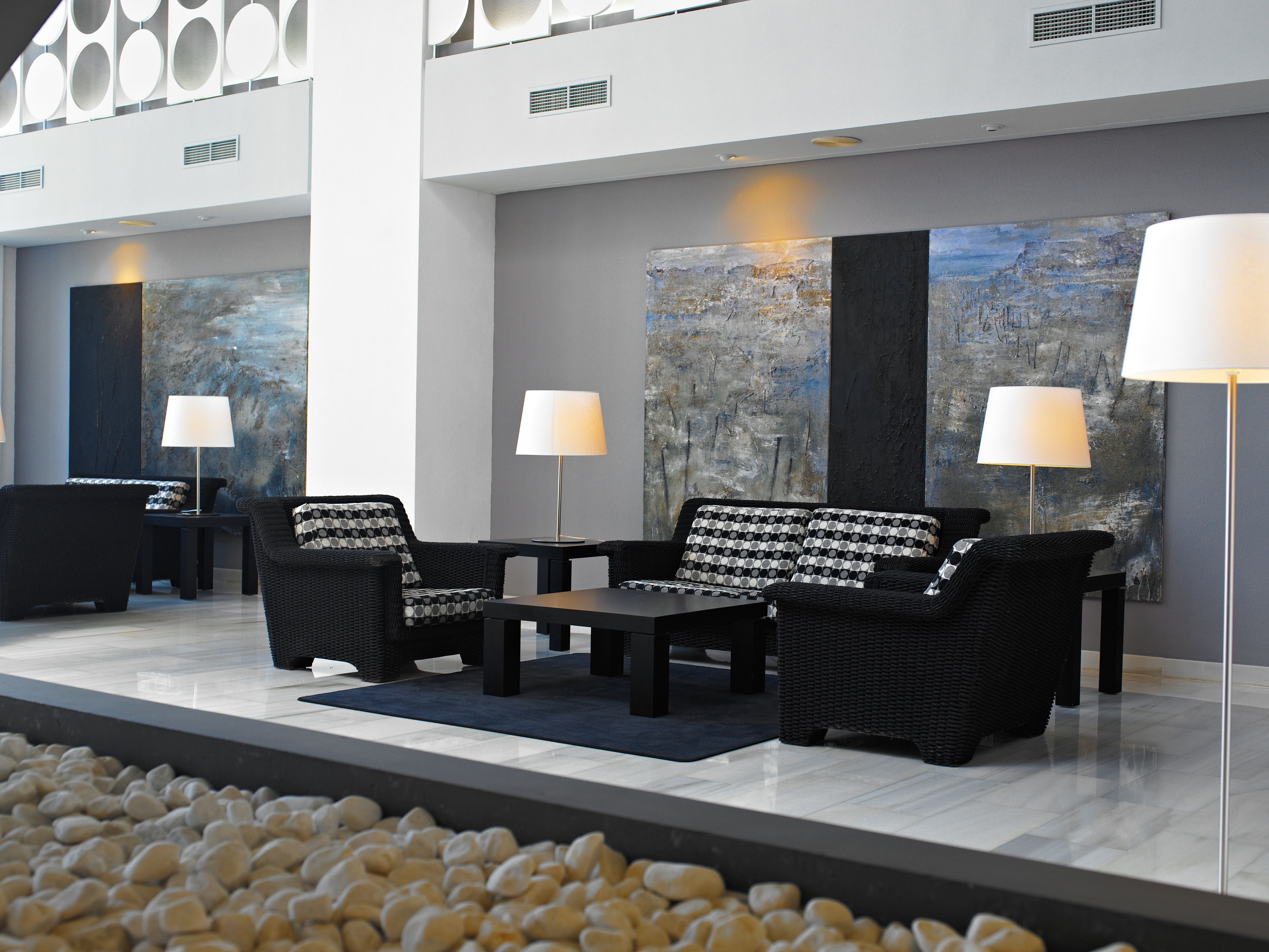 A modern lounge area with black wicker furniture, white lamps, and a large abstract painting on the wall.