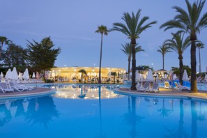 A pool area with lounge chairs and umbrellas, palm trees, and a building with lights in the background. - Protur Vista Badía Aparthotel (Sant Llorenc des Cardassar)
