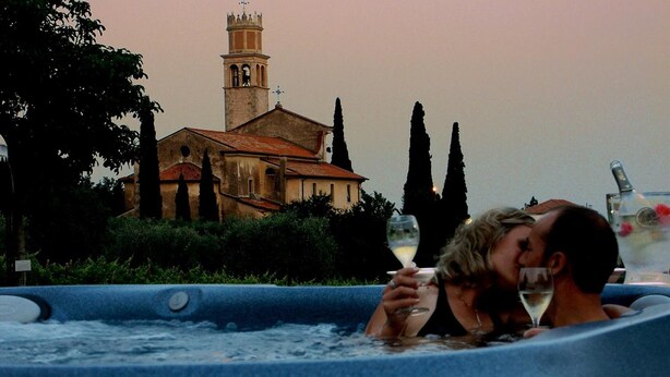 Un jacuzzi con una persona sosteniendo un vaso, una iglesia con campanario, y cipreses al fondo.