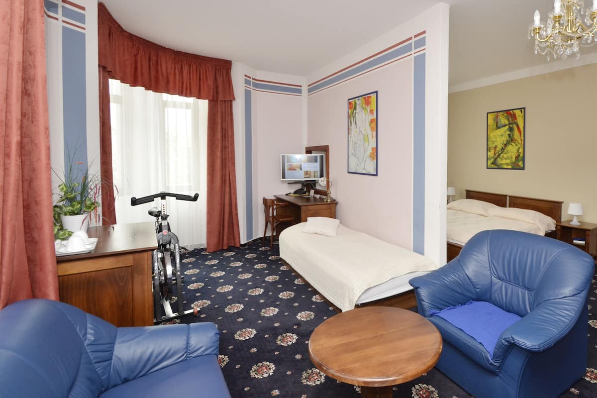 Une chambre d’hôtel avec un lit, deux fauteuils bleus, une petite table ronde, un bureau avec un vélo, une télévision et des œuvres d’art accrochées aux murs.