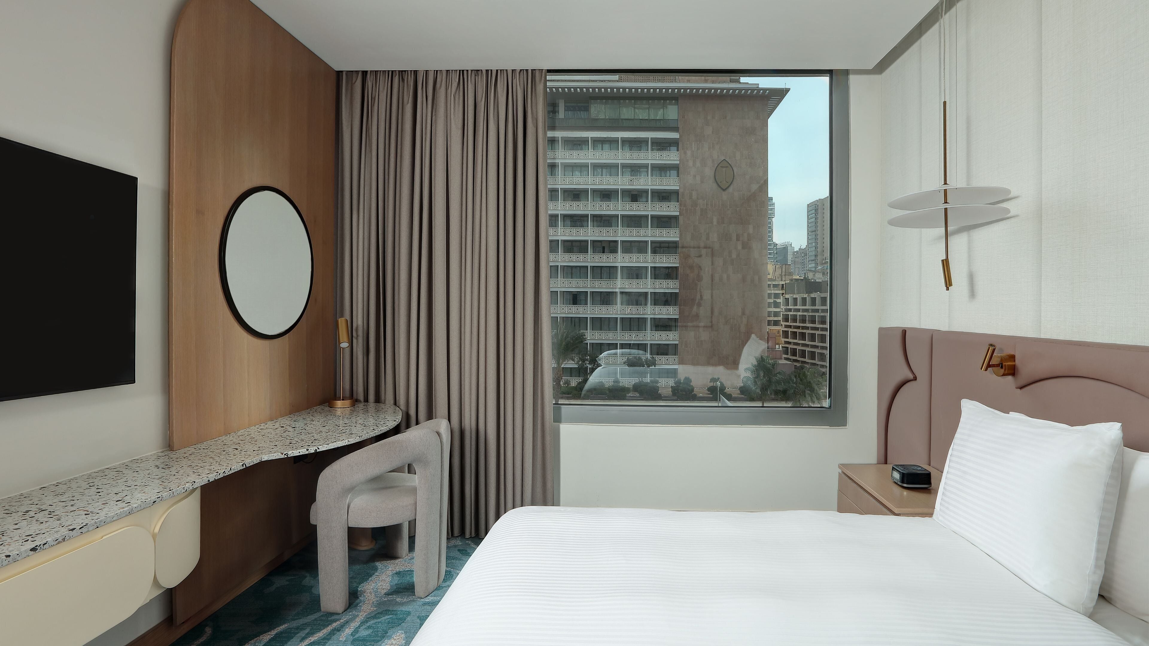 A hotel room with a bed, a desk, a chair, a TV, a window with a view of buildings, and a bedside table with a lamp.