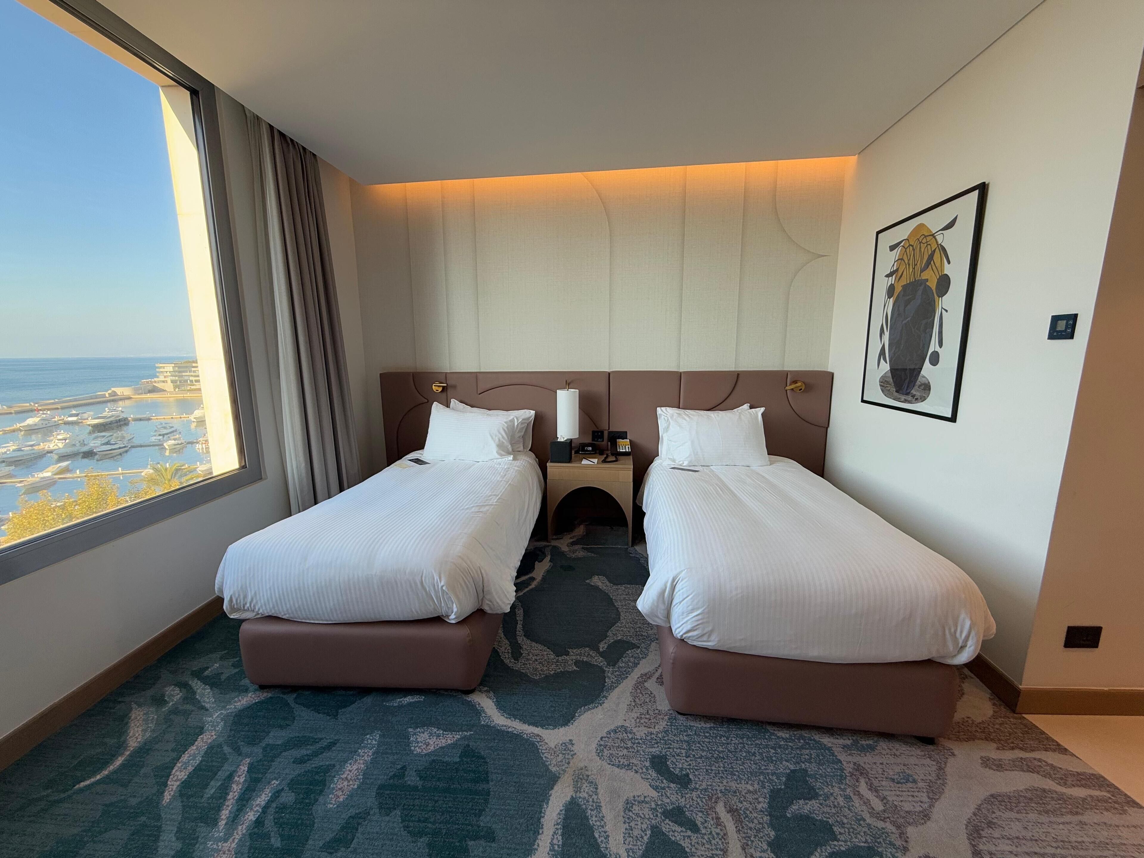 A hotel room with two beds, a view of the marina, and a painting on the wall.