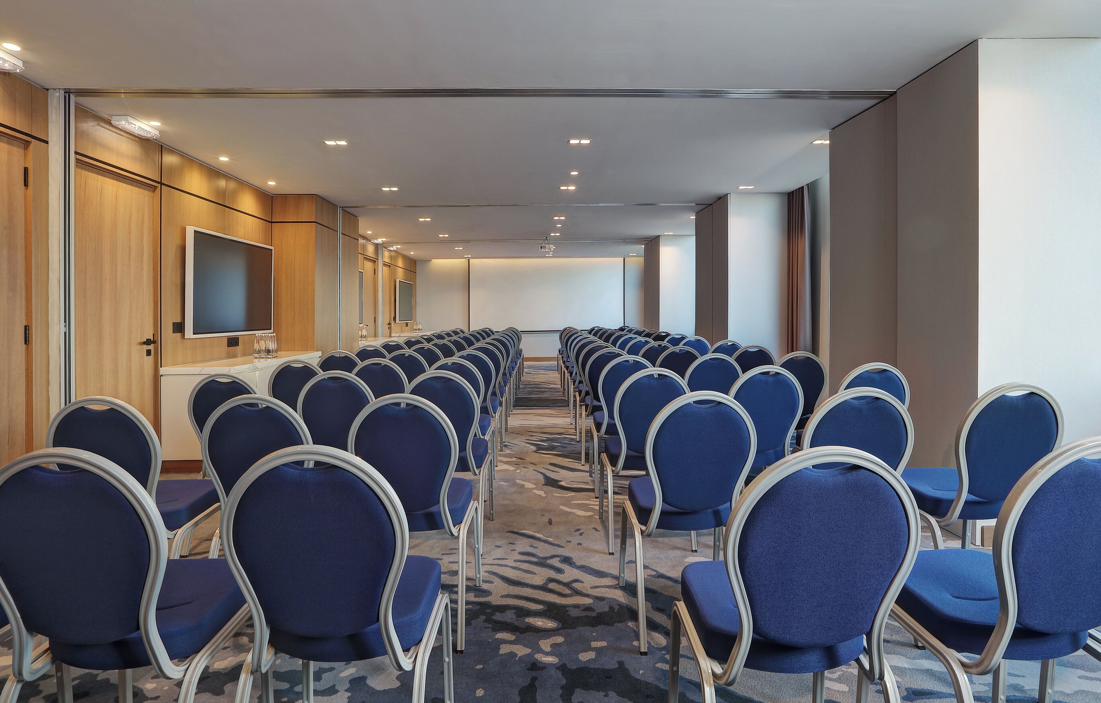 A conference room with rows of blue chairs, a large screen, and wooden paneling on the walls.