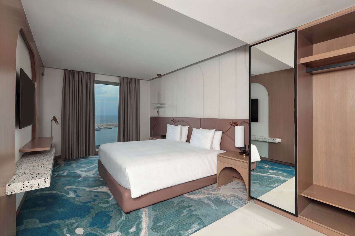 A hotel room with a large bed, a bedside table with a lamp, a TV mounted on the wall, and a view of the sea through the window.