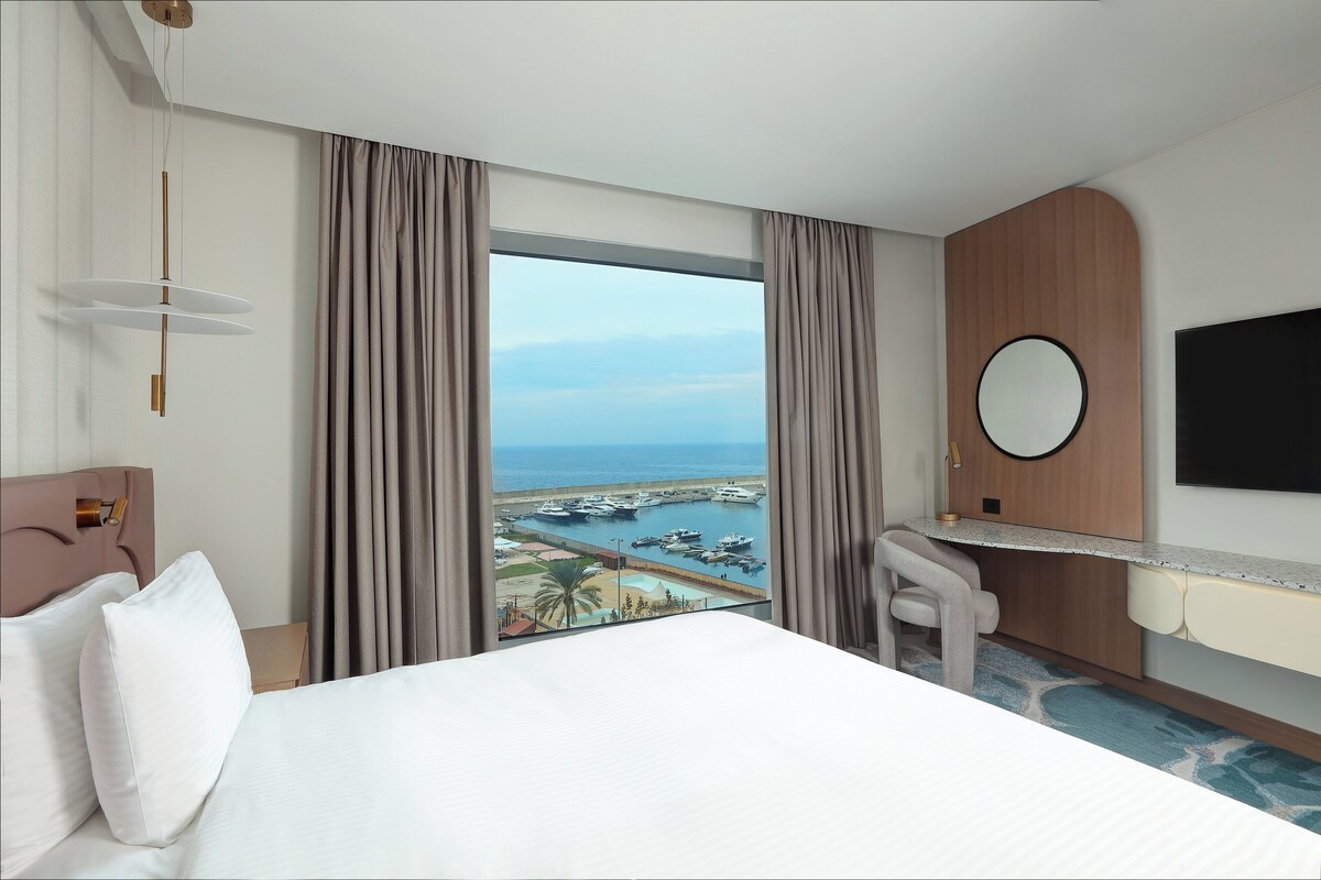 A hotel room with a bed, a desk, a chair, a TV, and a view of a marina.