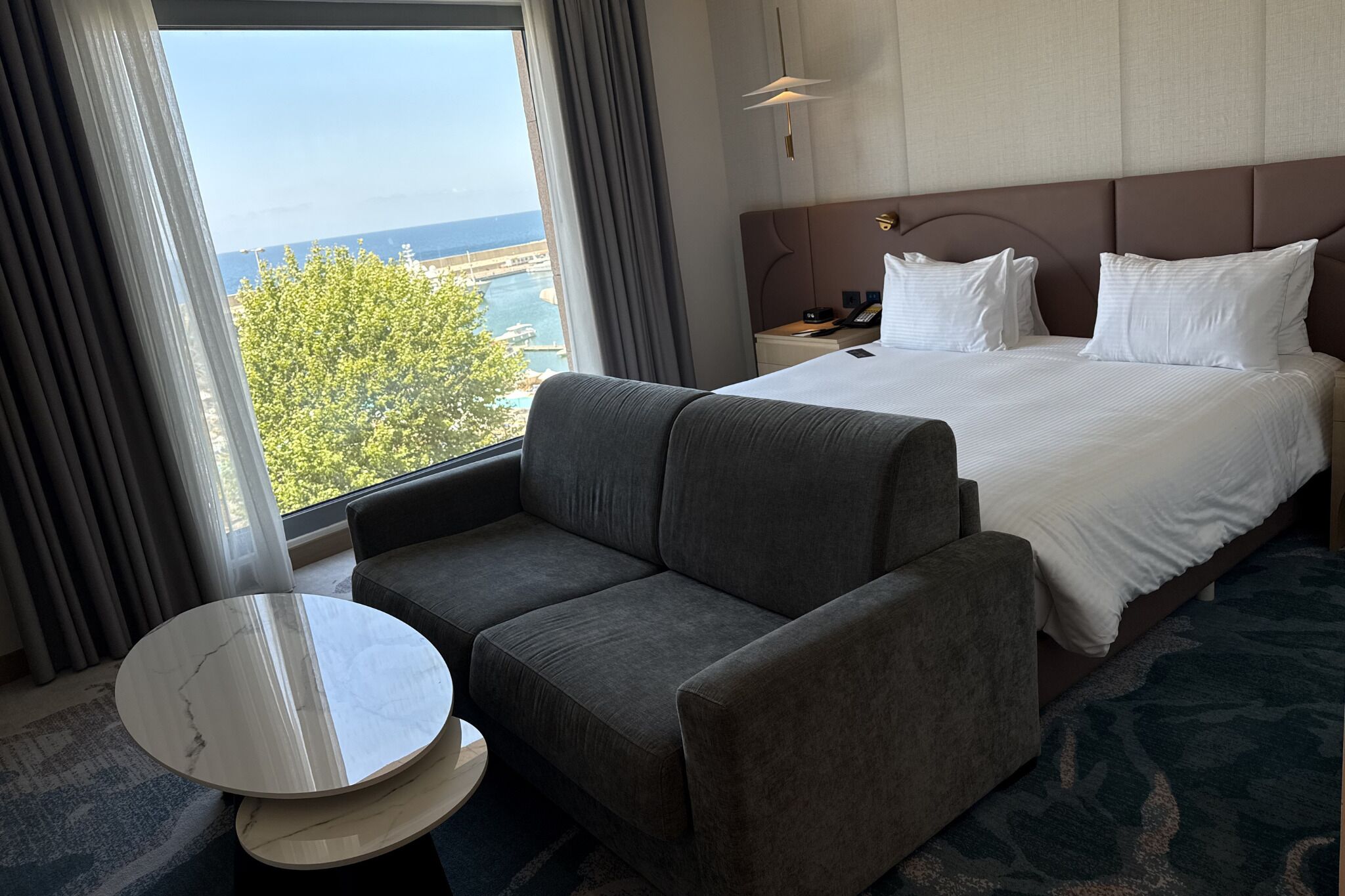 A hotel room with a bed, a sofa, a small table, and a view of the sea.