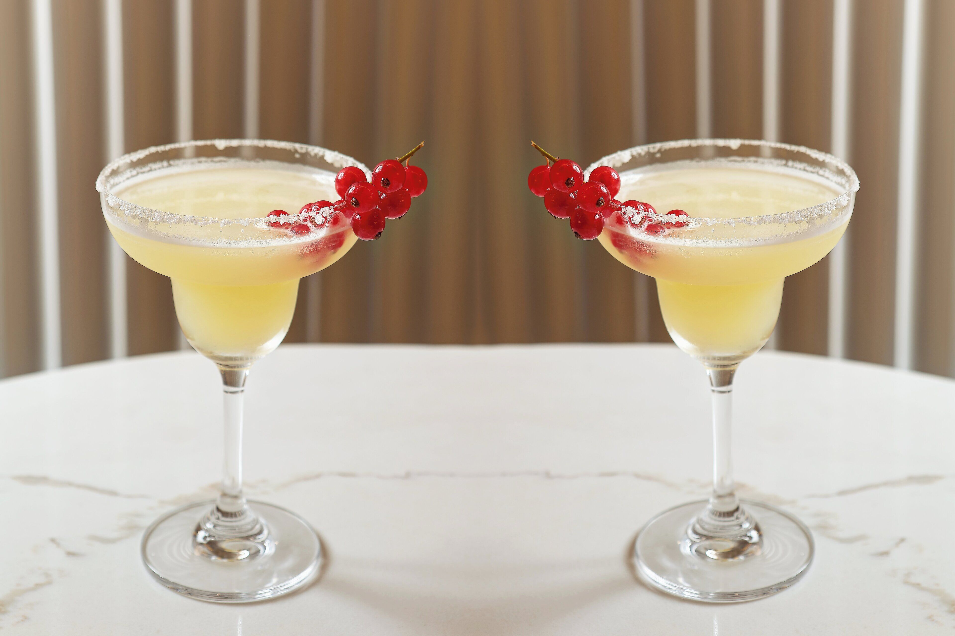 Twee martini glazen met een geel drankje en rode bessen op een witte tafel.
