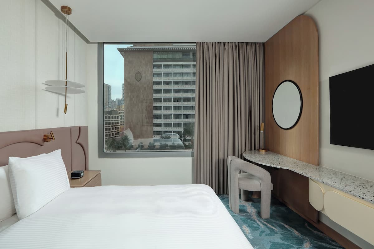 A hotel room with a large bed, a desk with a chair, a mirror, and a view of a cityscape through a window.