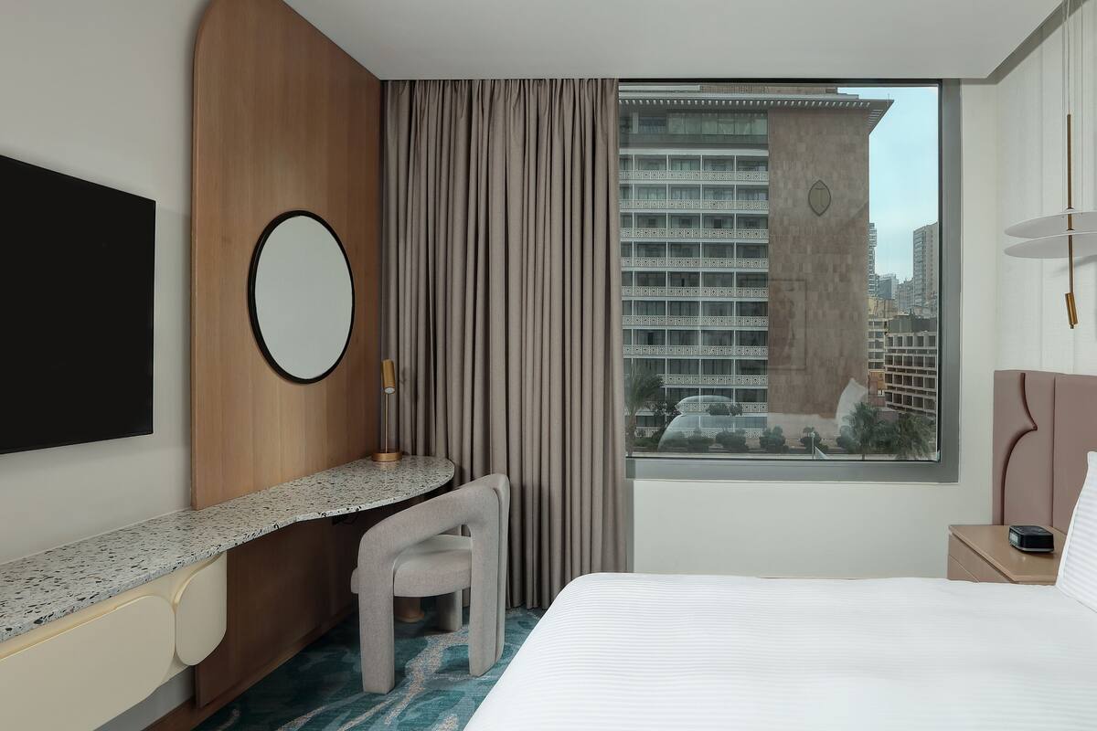 A hotel room with a bed, a desk, a chair, a TV, a window with a view of buildings, and a bedside table with a lamp.
