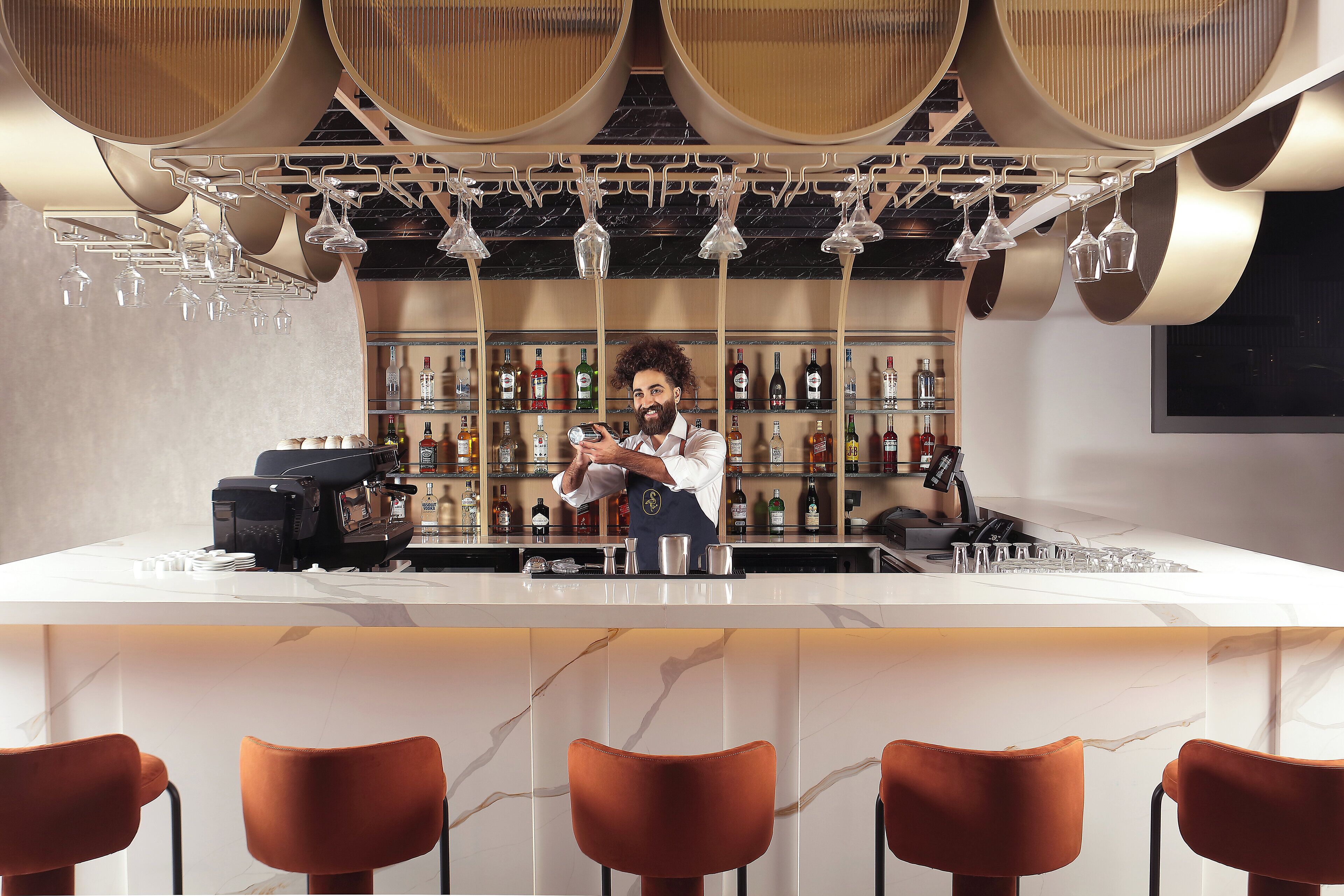 A bar area with a barista preparing drinks, a coffee machine, and a selection of bottles on shelves.