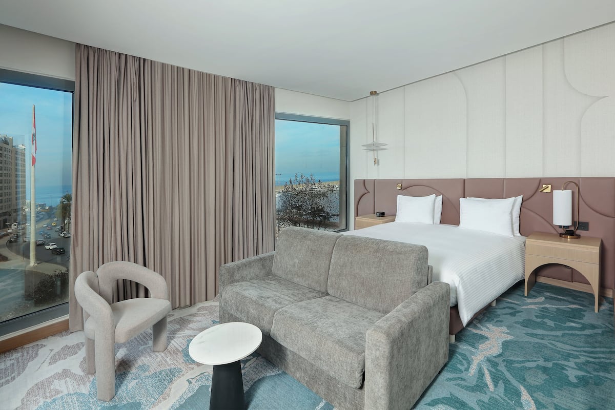 A modern hotel room with a large bed, a sofa, a small table, and a view of the city through a large window.