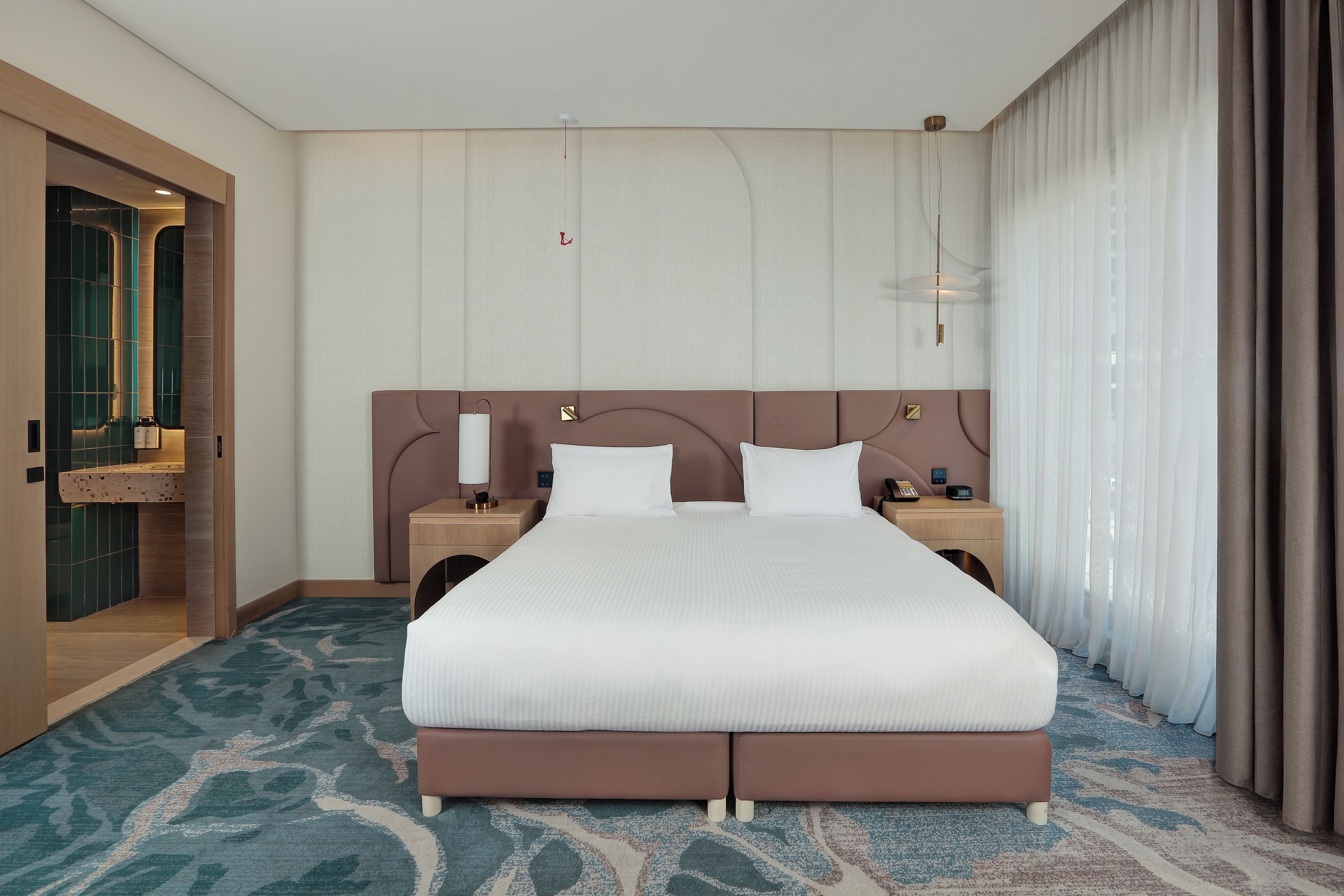 A hotel room with a large bed, a bedside table with a lamp, a nightstand, and a bathroom with a mirror and sink.