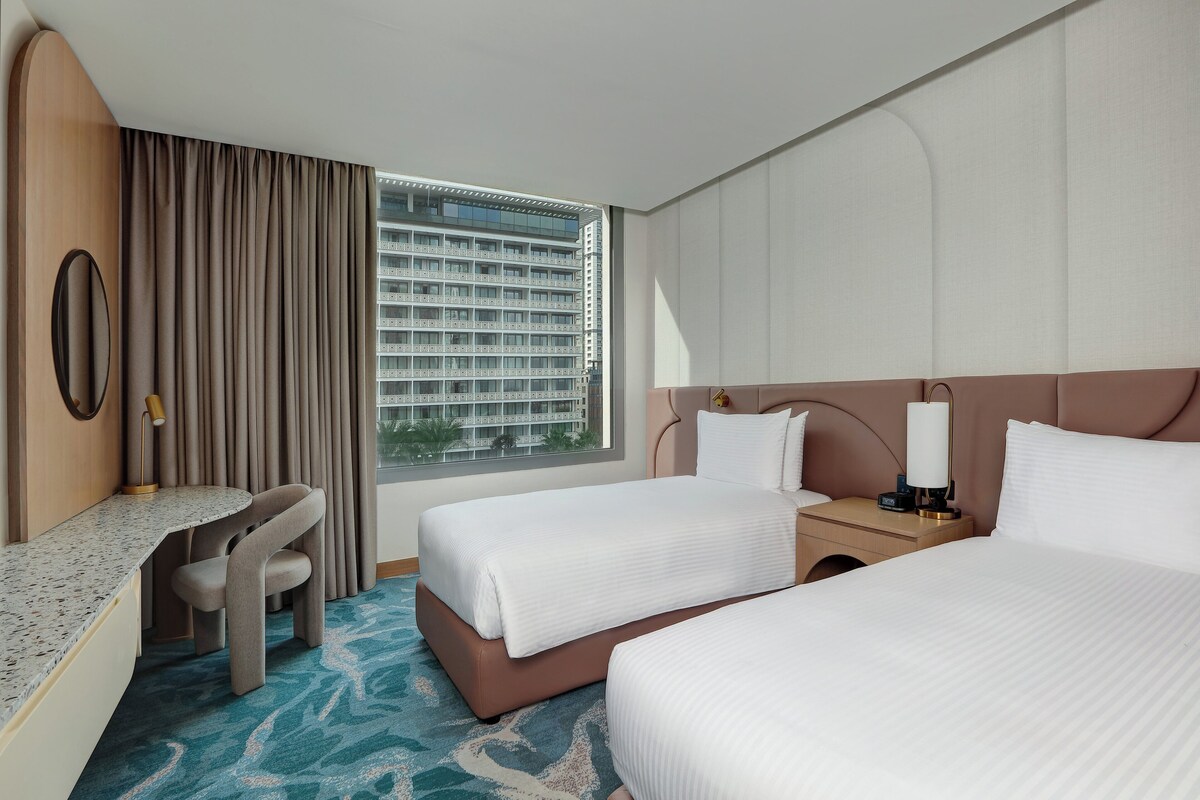 A hotel room with two beds, a desk, and a view of a high-rise building.