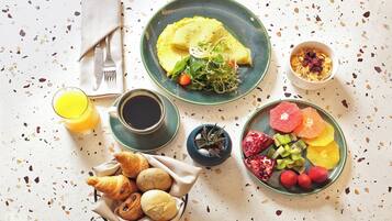 Un desayuno con huevos en un plato, cereal en un bol, un vaso de zumo de naranja, una taza de café, fruta en un plato y una cesta de cruasanes.