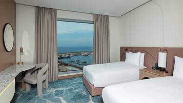 Habitación de hotel con dos camas, un escritorio, una silla y un ventanal con vistas a barcos y al agua.