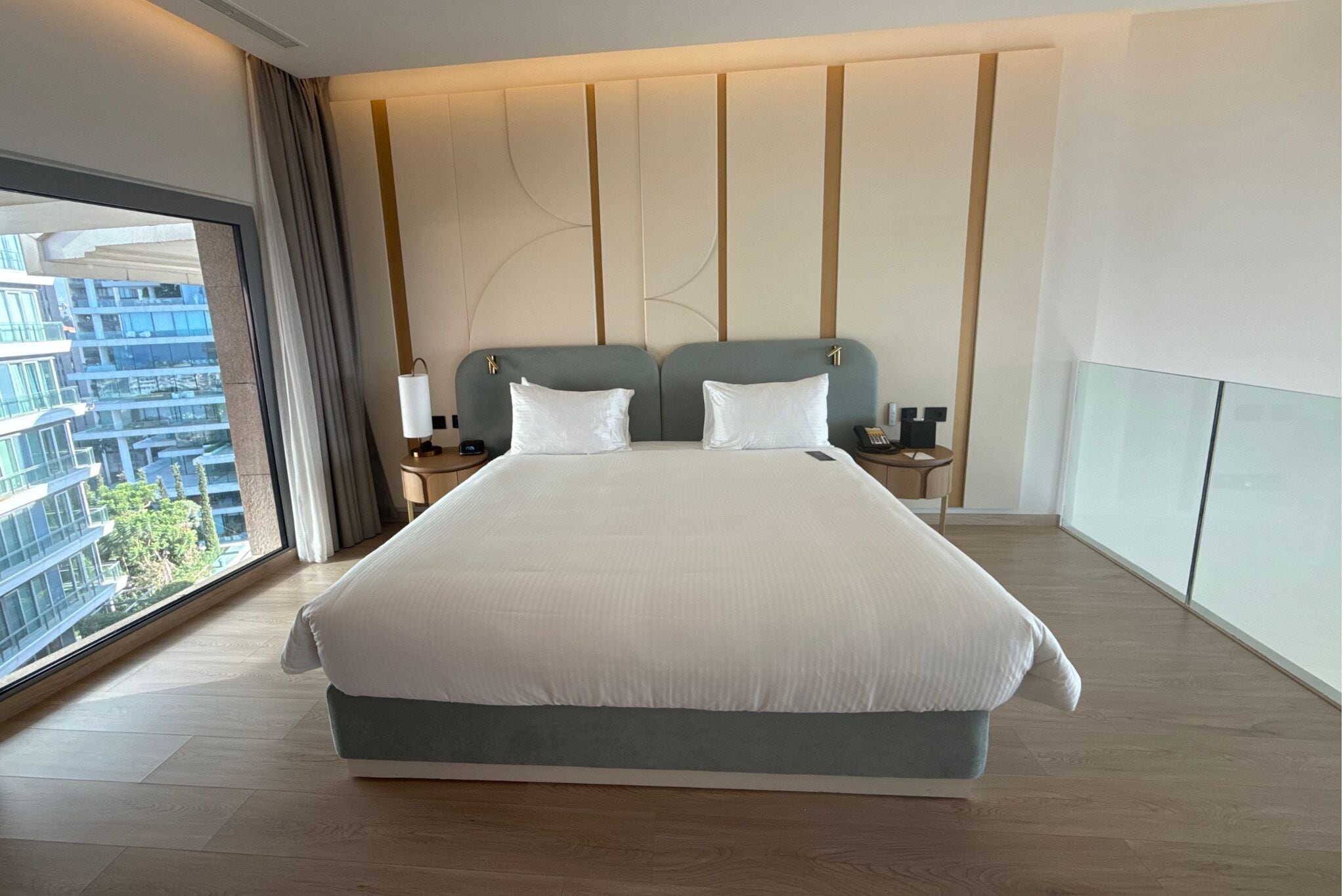 Een groot bed met twee kussens, een nachtkastje met een lamp, en uitzicht op een gebouw door een groot raam.