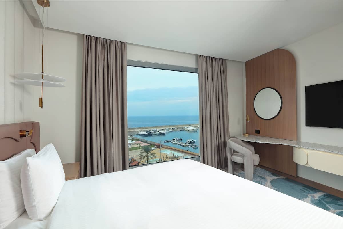 A hotel room with a bed, a desk, a chair, a TV, and a view of a marina.