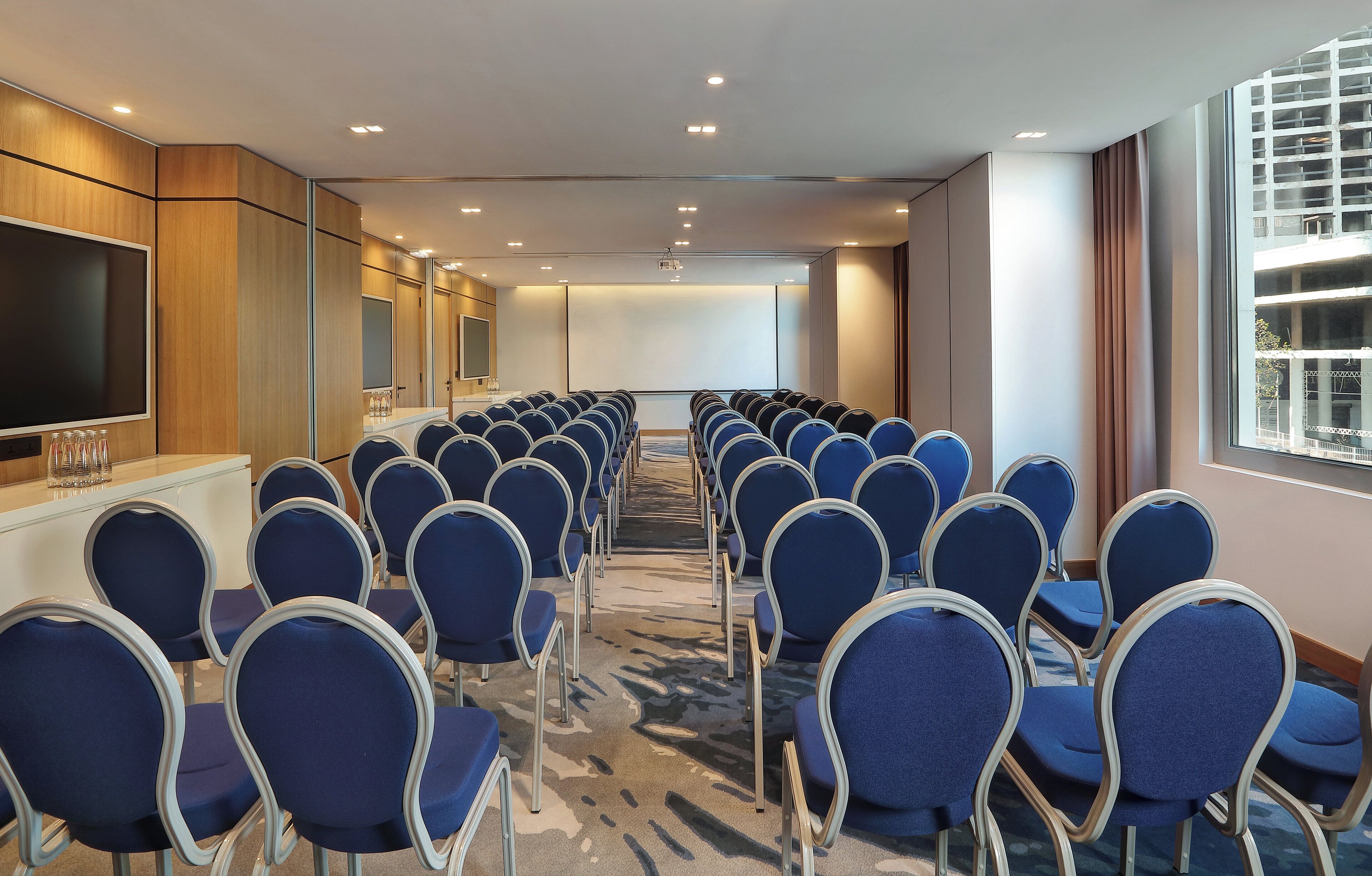 Un salón de conferencias con filas de sillas azules, una pantalla grande y suelo alfombrado.