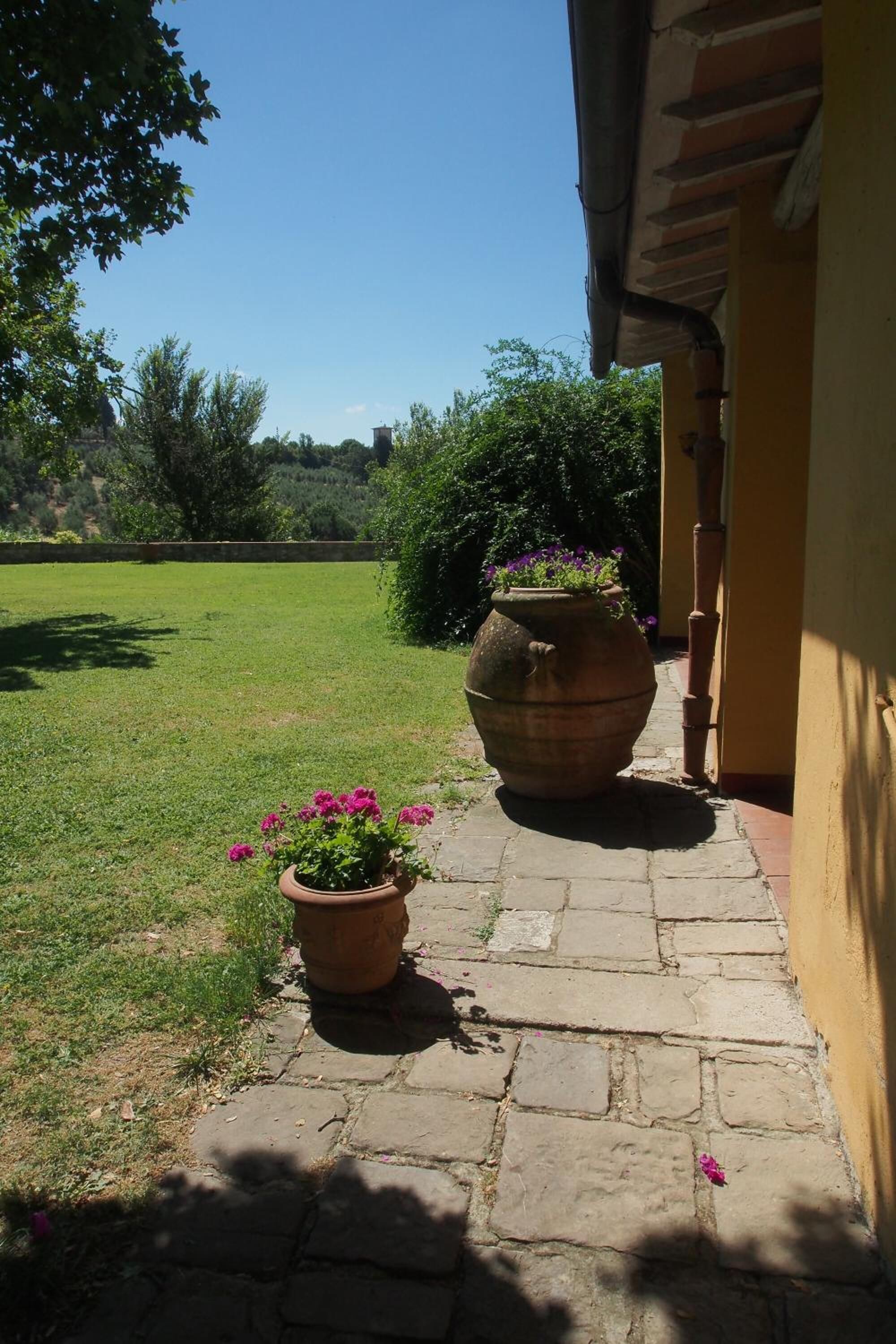 A pathway leading to a building with a yellow exterior, a large terracotta pot with flowers, and a grassy area in the background.