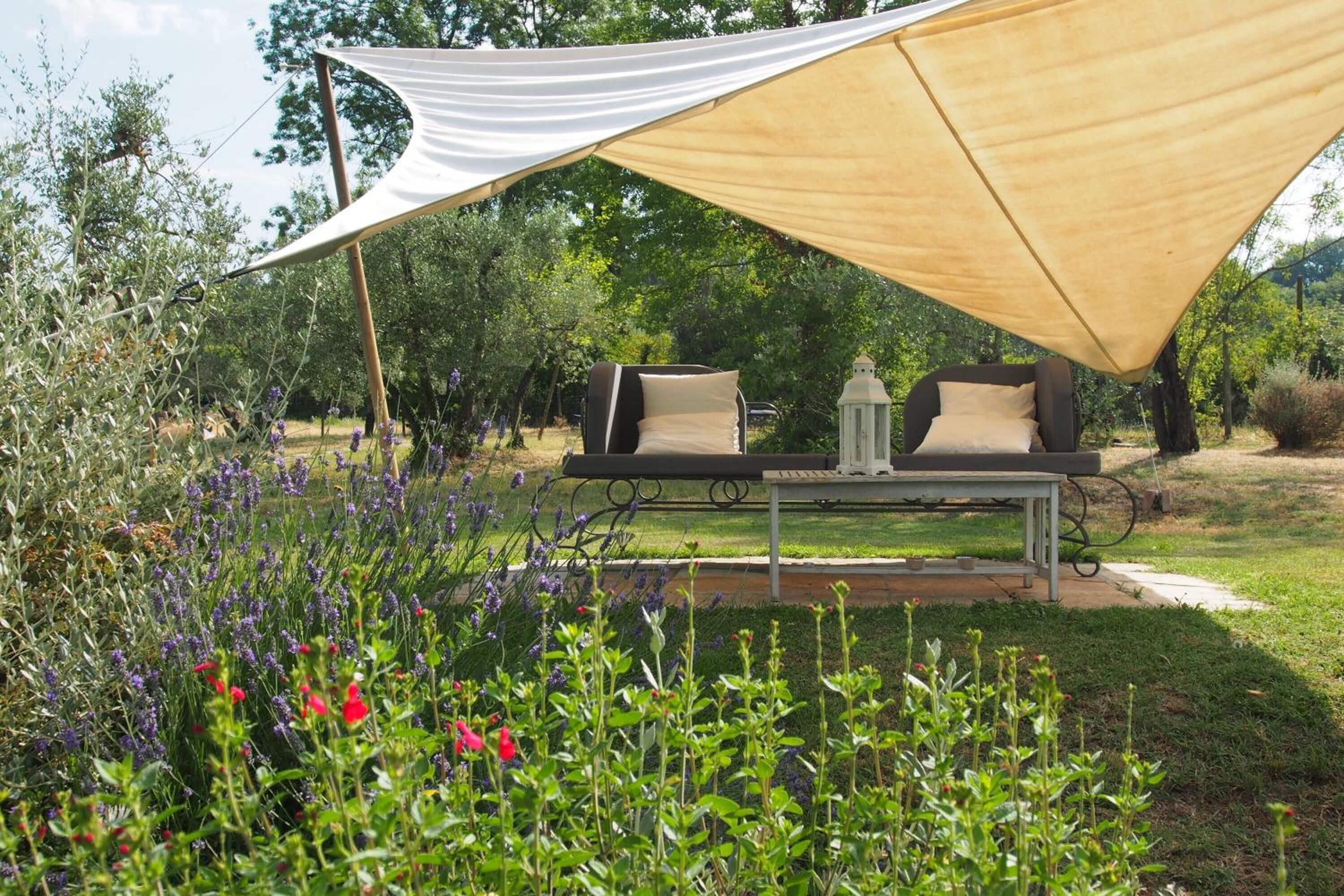 A garden with a table and chairs under a shade sail, surrounded by various plants and flowers.