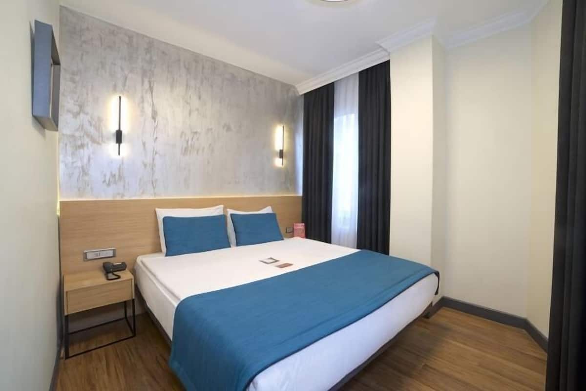 A hotel room with a large bed, a bedside table, a wall-mounted TV, and a window with curtains.
