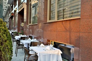 Un área de comedor al aire libre con mesas preparadas para dos, con sillas de mimbre y manteles blancos.