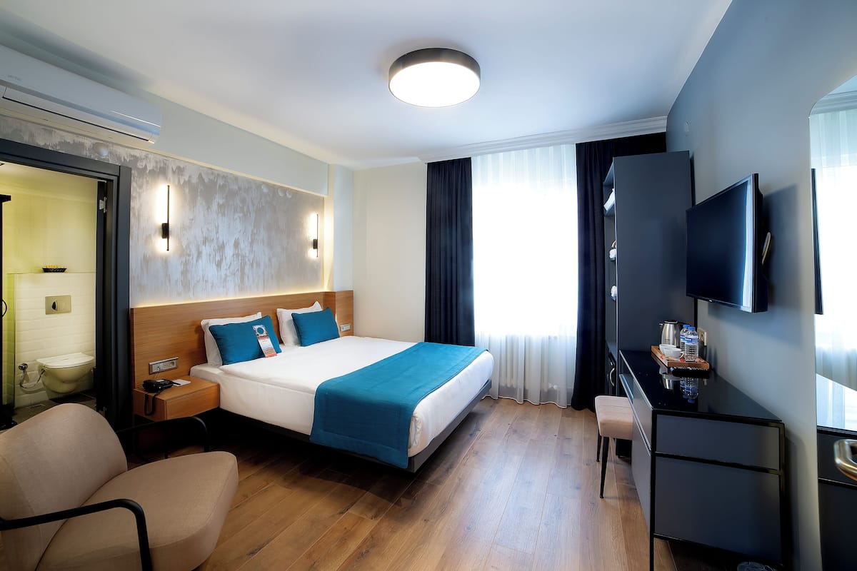 A hotel room with a bed, bedside table, chair, desk, TV, and a view of the outside.
