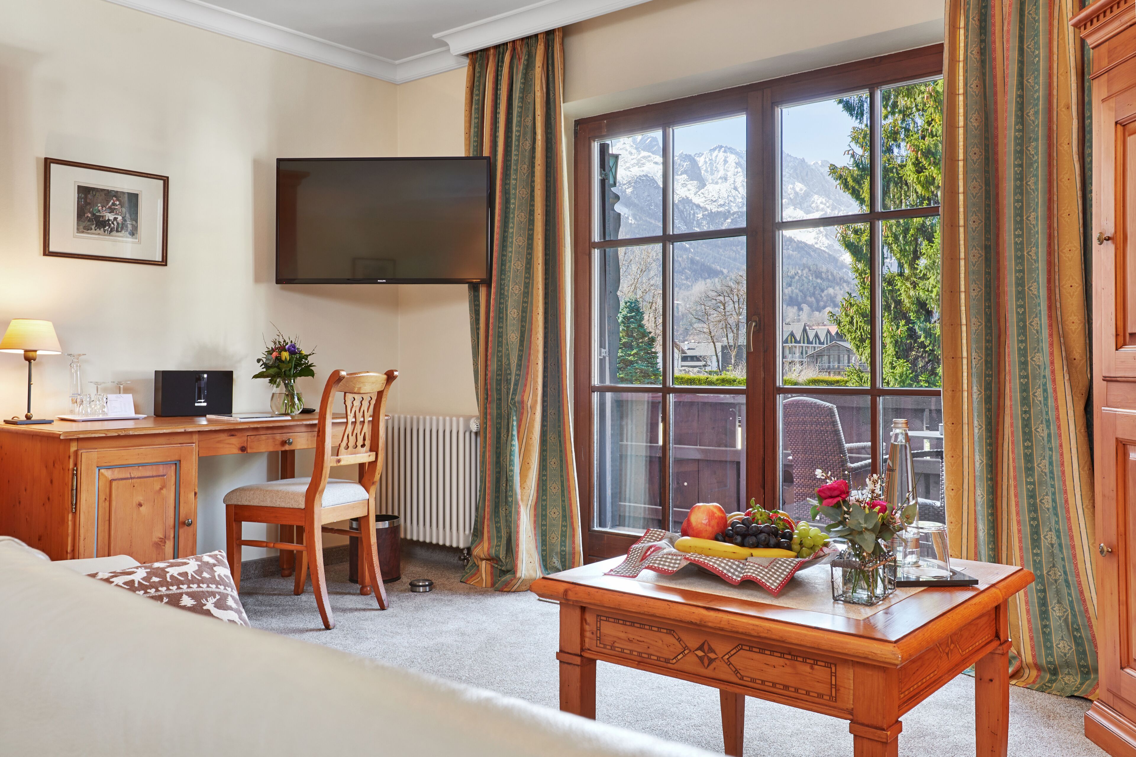 A hotel room with a wooden desk, a chair, a television, a large window with a mountain view, and a table with fruit and flowers.