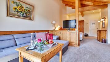 A living room with a sofa, a coffee table with flowers and fruit, a painting of sunflowers, wooden cabinets, a television, and a door leading to another room.