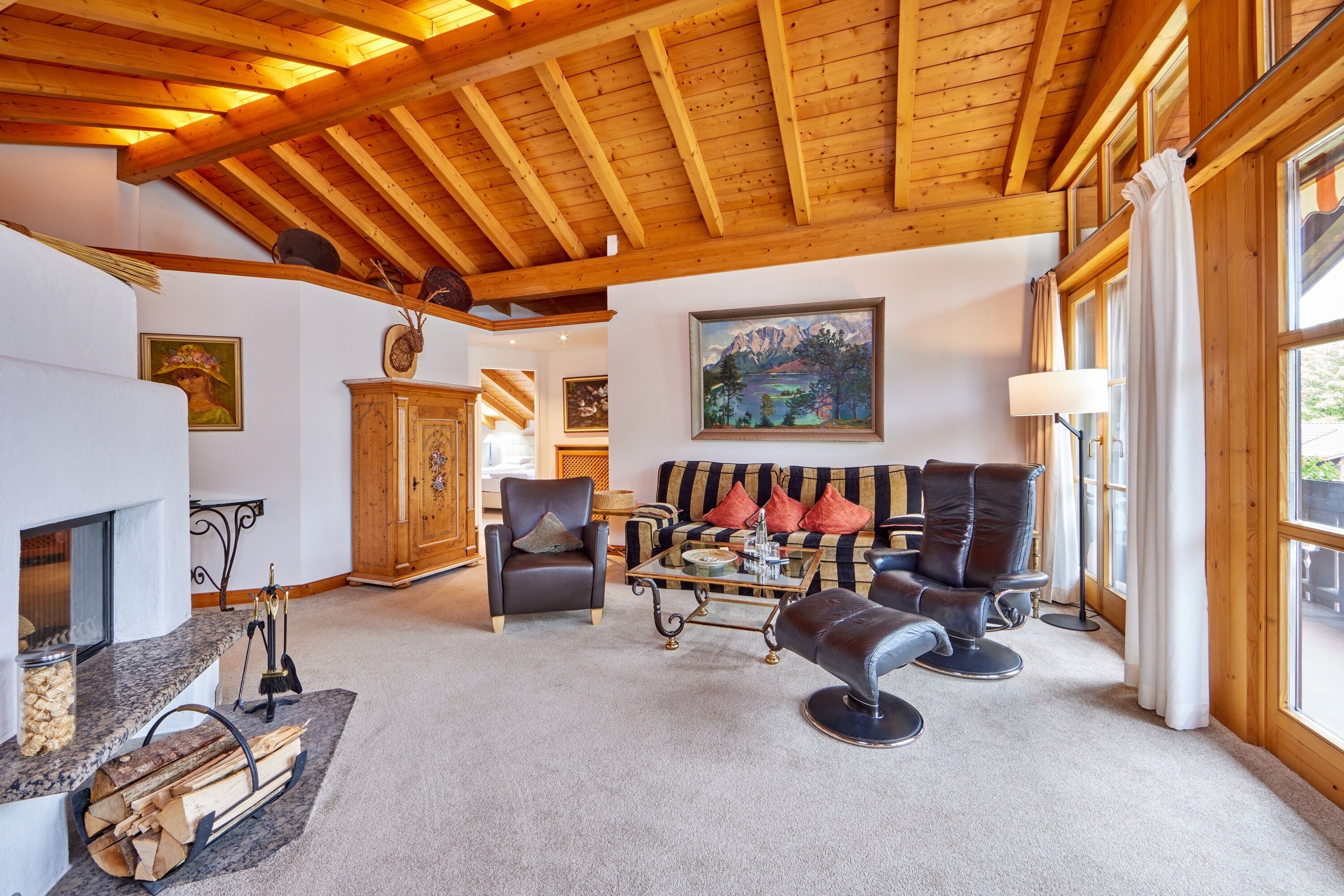 A cozy living room with a fireplace, wooden ceiling beams, a sofa with colorful cushions, and a painting of a lake.