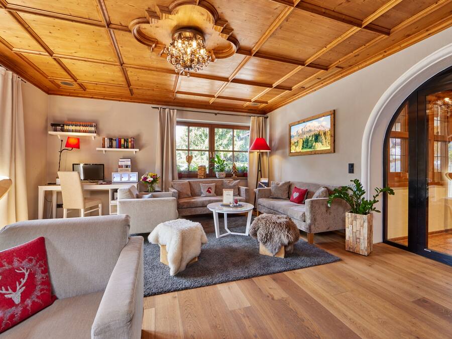 A cozy living room with a wooden ceiling, a chandelier, a fireplace, and a large window with a view of greenery.