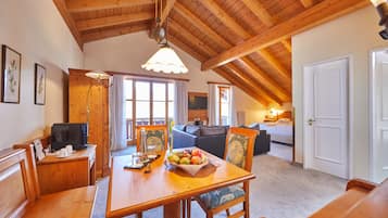 Un comedor con una mesa de madera, sillas y un frutero. Hay un televisor sobre un mueble de madera, un sofá al fondo y una puerta a la derecha.
