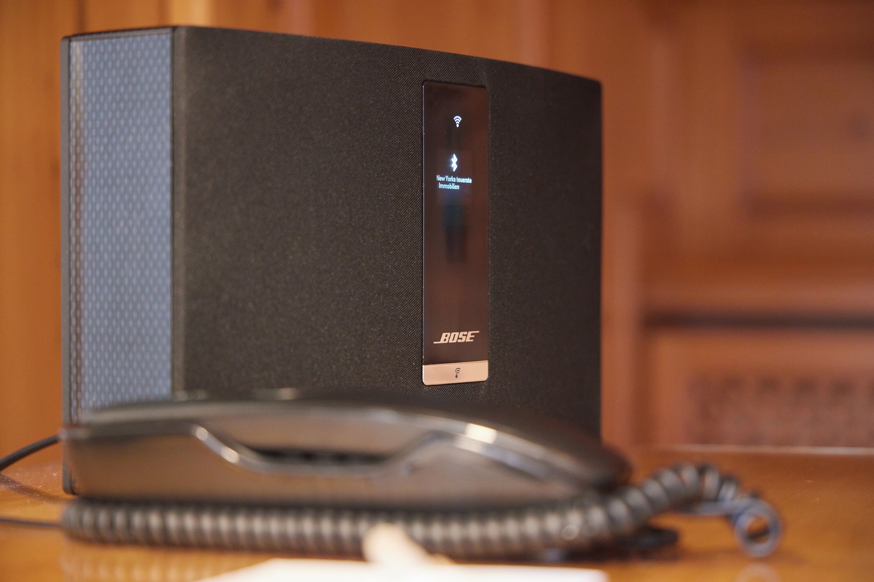 A black Bose speaker with a remote control and a coiled cord on a wooden surface.