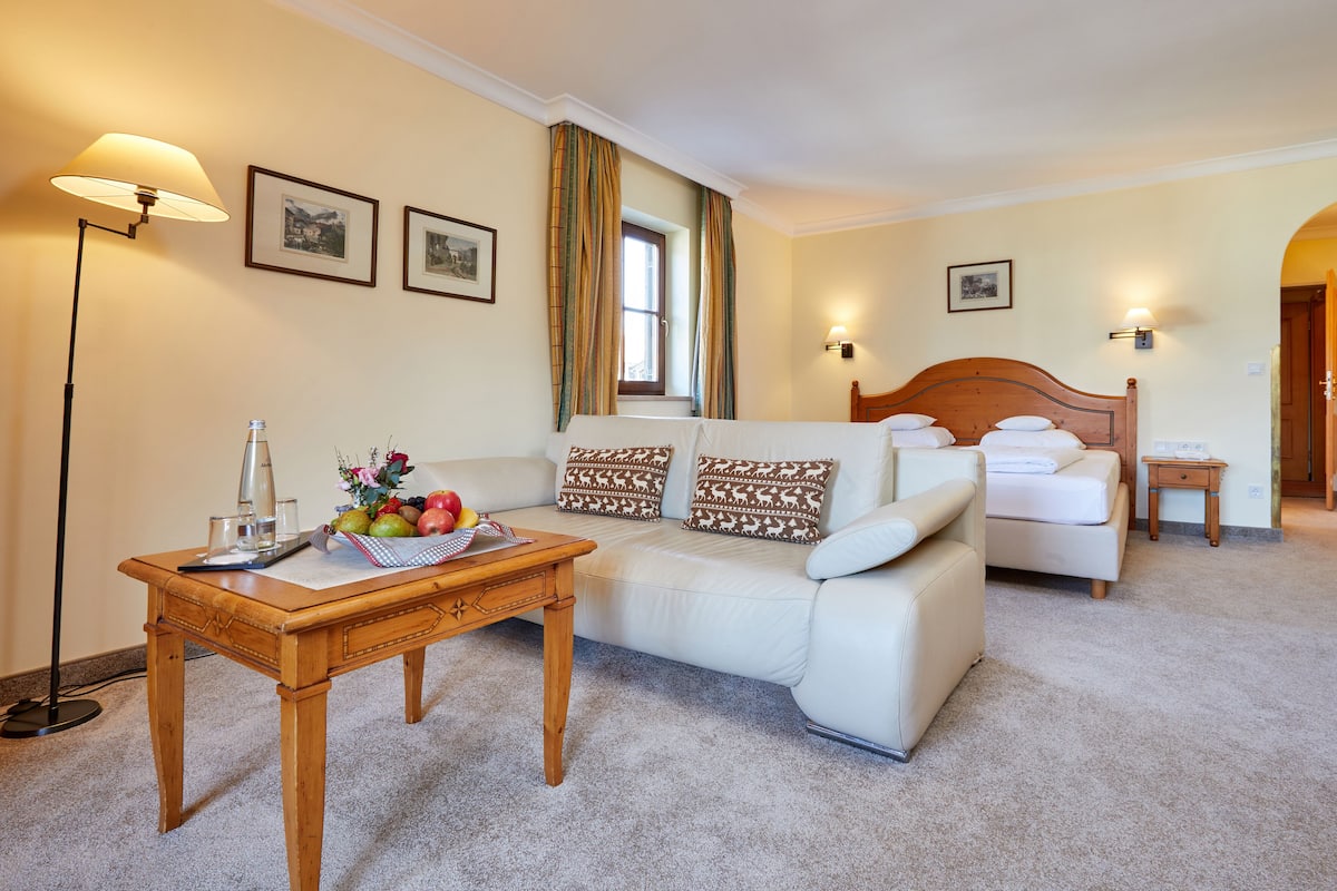 A hotel room with a sofa, a wooden table with a fruit bowl, a bed with white bedding, and two wall-mounted lamps.