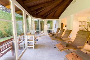 A sunlit room with large windows, wooden benches, and reclining chairs.