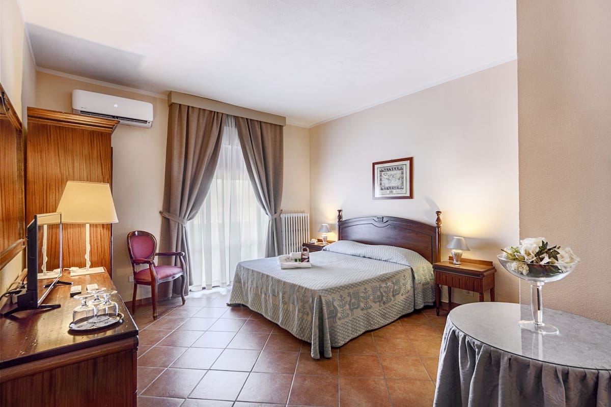 Una camera d'albergo con un letto, comodini, una scrivania con una lampada, una sedia, un tavolino con un vaso e un condizionatore d'aria.