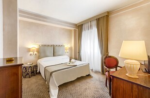 Habitación de hotel con cama, mesita de noche, lámpara, silla y cortinas.